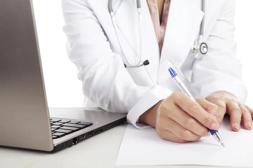 Close-up-of-medical-professional-writing-on-paper-looking-at-laptop