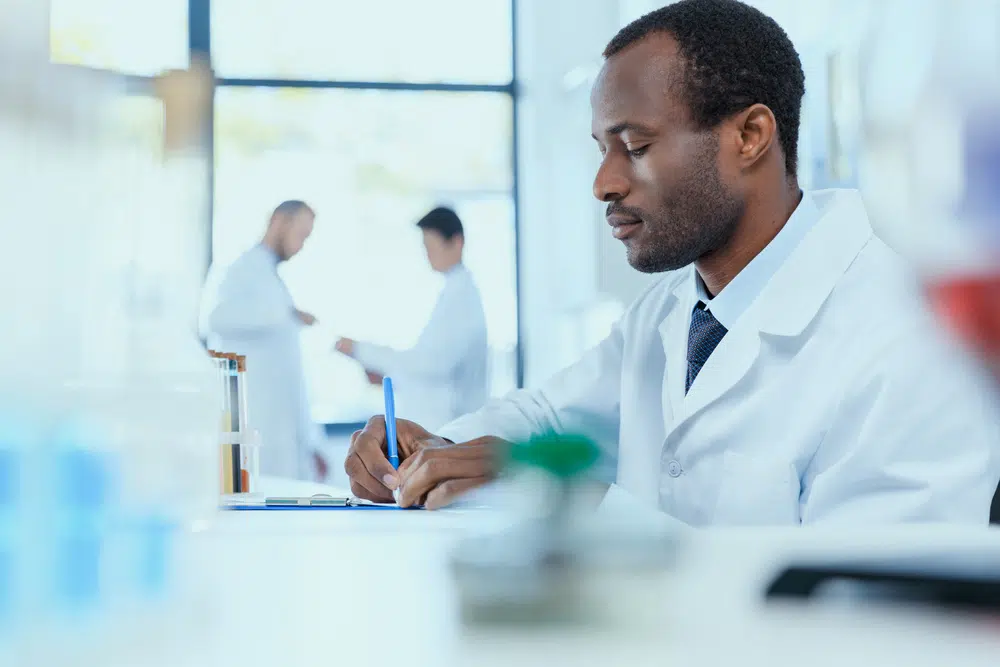 Young-man-in-lab-coat-writing-at-table-two-men-in-background
