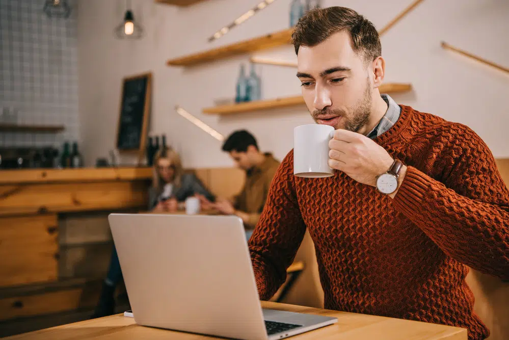 Young-man-in-sweater-at-coffee-shop-sipping-coffee-looking-at-laptop